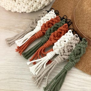 May include: Five macrame keychains with tassels in various colors, including white, beige, green, and orange. The keychains are hanging from a brown hat.