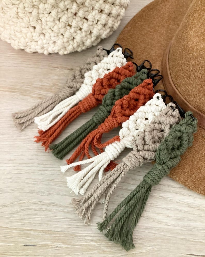 May include: Five macrame keychains with tassels in various colors, including white, beige, green, and orange. The keychains are hanging from a brown hat.
