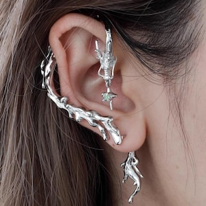 May include: Silver-toned ear jewelry set. The set includes a dragon ear cuff, a dragon-shaped earring with a green gem, and a dangling earring. The ear cuff has a branch-like design. The jewelry is worn on the ear.