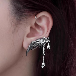 May include: A silver-toned ear cuff featuring a dark, leaf-like design that wraps around the ear. The cuff has dangling, decorative elements with a crystal-like appearance. The brand name "NOFEE" is visible at the bottom.