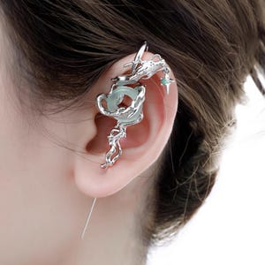 May include: Silver ear cuff featuring a dragon design with a light green stone and a star-shaped charm. The ear cuff wraps around the ear and has intricate details. The jewelry is worn on the ear.