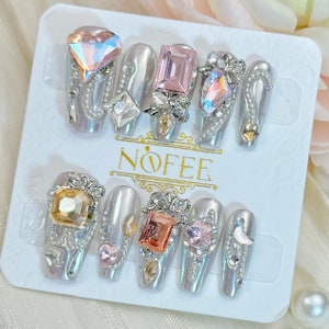 May include: A set of ten silver press-on nails with various embellishments, including rhinestones, pearls, and small charms. The nails are decorated with a variety of colors, including pink, peach, and gold. The nails are arranged on a white background with the word "NOFEE" printed in gold.