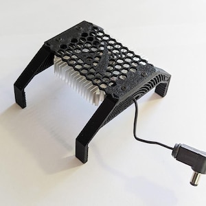 Puede incluir: Un soporte de enfriamiento impreso en 3D negro con un diseño de panal y un disipador de calor plateado. El soporte tiene un cable de alimentación negro con un enchufe en ángulo recto.