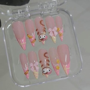Op de afbeelding: Een set van tien kunstnagels in een doorzichtige plastic doos. De nagels zijn voornamelijk roze met verschillende ontwerpen, waaronder strikken, stripfiguren en gele accenten. Sommige nagels hebben een French tip-ontwerp met roze en gele details.