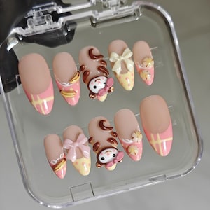 Könnte beinhalten: Ein Set aus zehn künstlichen Nägeln in einer transparenten Kunststoffbox. Die Nägel weisen eine Vielzahl von Designs auf, darunter einfarbige Nude-, Rosa- und Gelbmuster, Schleifen und Zeichentrickfiguren. Die Nägel sind oval und haben ein glänzendes Finish.