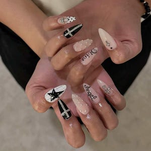 Puede incluir: Un conjunto de uñas acrílicas largas y puntiagudas con varios diseños. Algunas uñas son negras con cruces blancas, otras blancas con estrellas negras y algunas nude con pedrería. Las palabras "Divine" y "Angel" están escritas en dos uñas.
