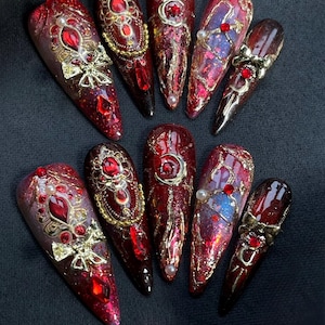 Könnte beinhalten: Ein Set aus zehn stilettoförmigen Press-on-Nägeln mit einer dunkelroten Glitzerbasis. Die Designs umfassen goldene Metallschleifen, verzierte Filigranrahmen, kleine weiße Perlen und eingebettete rote Strasssteine in Mustern.