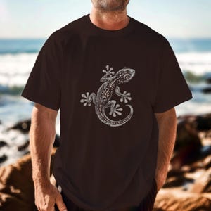 Könnte beinhalten: Ein dunkelbraunes T-Shirt mit einer detaillierten weißen Illustration eines Geckos. Das Gecko-Design ist mittig platziert und weist komplizierte Muster auf. Das T-Shirt hat kurze Ärmel und einen Rundhalsausschnitt.
