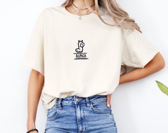 May include: Cream-colored t-shirt featuring a small black illustration of an alpaca with the word "ALPACA" underneath. The shirt has short sleeves and a relaxed fit, suitable for casual wear. The design is simple and minimalist.