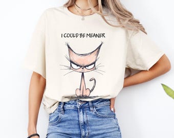 May include: Cream-colored t-shirt featuring a grumpy cat illustration with the text "I COULD BE MEANER" in black. The cat has an angry expression with a pink body and black whiskers. The shirt is made of a soft material.