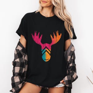 May include: Black t-shirt featuring a colorful moose head graphic. The moose antlers are pink and orange, with a teal and orange chevron design below. The shirt is worn with a black and gray plaid shirt.