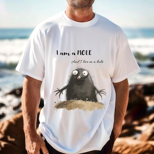 Könnte beinhalten: Weißes T-Shirt mit einem Cartoon-Maulwurf-Design. Der Maulwurf ist schwarz mit großen Augen und ausgestreckten Armen. Der Text über dem Maulwurf lautet "I am a MOLE" und "And I live in a hole."
