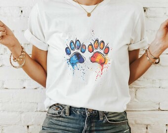 May include: White t-shirt with a colourful paw print design. The design features two paw prints, one blue and one orange, with paint splatter effects. The shirt is made of a soft material, suitable for casual wear.