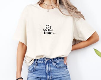 May include: Cream-colored t-shirt featuring a small black and white illustration of a rhino sitting in grass. The word "RHINO" is printed in black below the rhino graphic. The shirt is worn by a person in blue jeans.