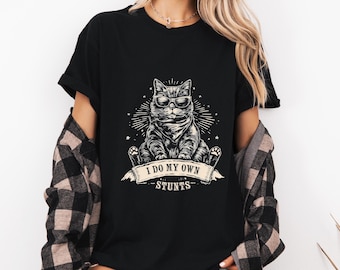 May include: Black t-shirt featuring a cool cat wearing sunglasses, with the text "I DO MY OWN STUNTS" on a banner. The cat graphic is surrounded by a starburst design. The shirt is worn with a plaid flannel shirt.