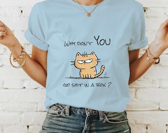 May include: Light blue t-shirt featuring a cartoon cat with an angry expression. The text on the shirt reads "WHY DON'T YOU GO SHIT IN A BOX?" The cat is orange with black outlines, and the text is in black.