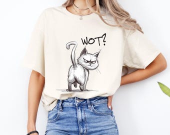 May include: Cream-colored t-shirt featuring a black and white illustration of a grumpy cat with the text "WOT?" above it. The cat is drawn in a cartoon style, with its tail curled and its expression conveying annoyance.