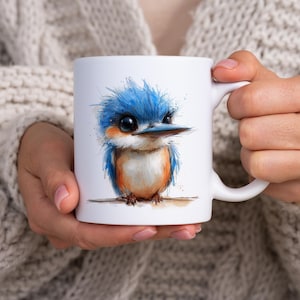 Op de afbeelding: Witte keramische mok met een aquarel illustratie van een blauwe en oranje vogel. De vogel heeft grote ogen en zit op een tak. De mok wordt vastgehouden door een persoon die een crèmekleurige trui draagt. Een charmant en grillig ontwerp.