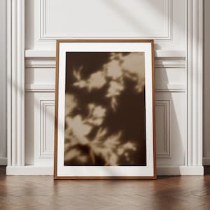 May include: A framed art print featuring a sepia-toned floral design. The artwork showcases the silhouette of flowers and leaves against a dark brown background. The frame is made of wood and is rectangular.