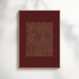 Può includere: Una stampa artistica astratta incorniciata. L'opera presenta un disegno dorato a forma di labirinto su uno sfondo rosso scuro. La cornice è di colore marrone chiaro. La stampa è esposta su una parete bianca.