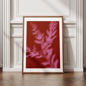 May include: A framed art print featuring a deep red background with a soft, blurred pink botanical design. The artwork is displayed in a wooden frame, leaning against a white wall and a wooden floor.
