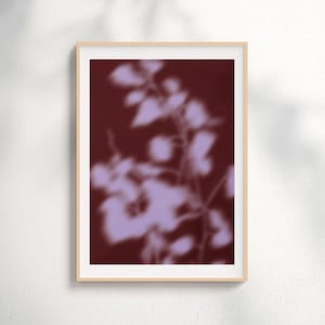 Op de afbeelding: Een ingelijste kunstdruk met een wazige afbeelding van een plant met lichtpaarse bladeren tegen een donkerrode achtergrond. Het kunstwerk zit in een lichtbruine houten lijst, ideaal voor wanddecoratie.
