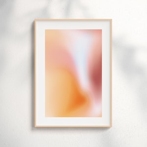 May include: A framed abstract art print featuring a soft, blurred gradient of colors. The artwork showcases a blend of warm hues, including peach, orange, and a touch of red, with a subtle white and blue accent. The print is encased in a light-colored wooden frame.