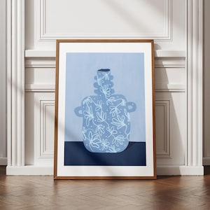 May include: A framed art print featuring a stylized blue vase with white floral patterns. The vase has two handles and a textured neck. The background is a light blue, with a dark blue base. The frame is a warm brown.