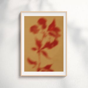Op de afbeelding: Een ingelijste kunstdruk met een wazige afbeelding van een rode bloemarrangement tegen een bruinachtige achtergrond. Het kunstwerk is ingelijst in een licht houten frame, wat een minimalistische uitstraling creëert. De afbeelding is zacht van focus, wat het een dromerige kwaliteit geeft.