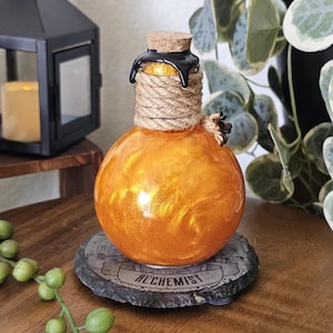 May include: An orange, spherical glass bottle with a cork stopper and rope detailing. The bottle sits on a stone coaster with the word "ALCHEMIST" inscribed. A black lantern and greenery are in the background.