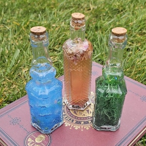 May include: Three decorative glass bottles with cork stoppers, each filled with a different colored liquid and decorative elements. The bottles are arranged on a maroon book against a backdrop of green grass.