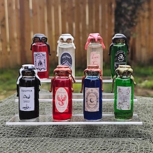 May include: Eight small glass bottles with cork stoppers and labels. The bottles contain colorful liquids and have labels with various text, including "Forgotten Souls", "Phoenix", "Love Tonic", and "Willow Water".