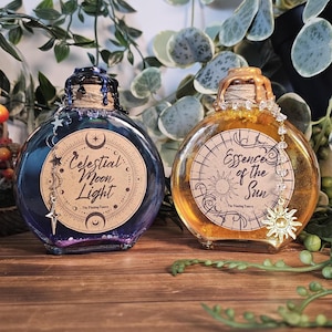 May include: Two round glass bottles with cork stoppers. One bottle contains blue liquid and is labeled "Celestial Moon Light." The other contains yellow liquid and is labeled "Essence of the Sun." Both have decorative charms.