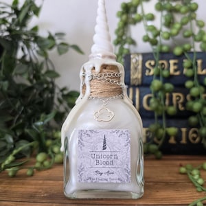 May include: A clear glass bottle filled with a shimmering white liquid, topped with a white spiral unicorn horn. The neck is wrapped in twine and a silver chain with a cloud charm. A label reads Unicorn Blood, Stay Alive, The Floating Tavern.