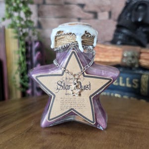 May include: A star-shaped glass bottle filled with a purple liquid, featuring a tan label with the text Star Dust. The bottle neck is wrapped in twine and sealed with white wax, and a silver chain with a small star charm hangs around it.