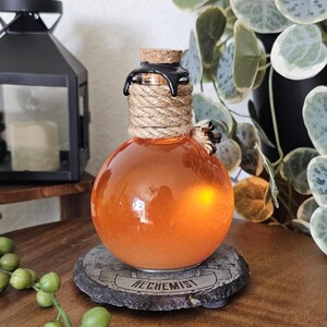 May include: A round, orange glass bottle with a cork stopper, wrapped in rope and sealed with black wax. The bottle sits on a stone coaster with the word "ALCHEMIST" inscribed. The bottle is illuminated from within.