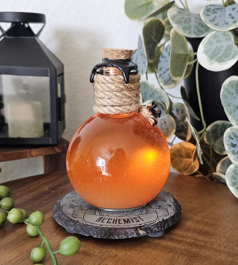 May include: A round, orange glass bottle with a cork stopper, wrapped in rope and sealed with black wax. The bottle sits on a stone coaster with the word "ALCHEMIST" inscribed. The bottle is illuminated from within.