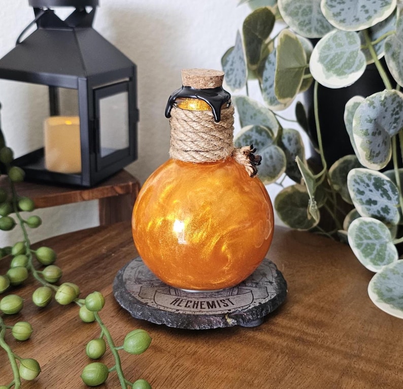 May include: An orange, spherical glass bottle with a cork stopper and rope detailing. The bottle sits on a stone coaster with the word "ALCHEMIST" inscribed. A black lantern and greenery are in the background.