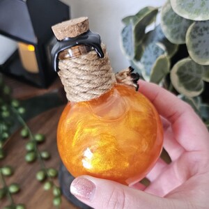 May include: A round, orange glass bottle with a cork stopper, wrapped with rope and sealed with black wax. The bottle contains a shimmering, golden liquid. Decorative item.