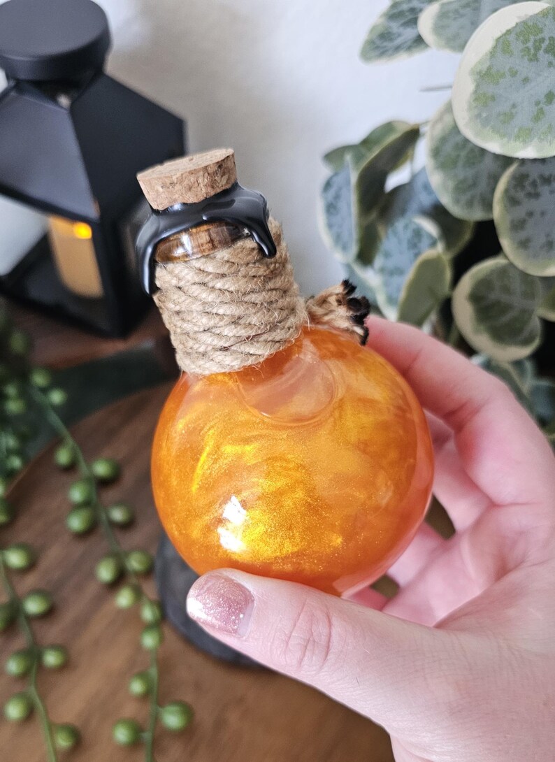 May include: A round, orange glass bottle with a cork stopper, wrapped with rope and sealed with black wax. The bottle contains a shimmering, golden liquid. Decorative item.