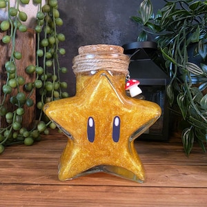 May include: A star-shaped glass bottle filled with gold glitter liquid. The bottle features two dark purple oval eyes on the front, a cork stopper wrapped in twine, and a small red and white mushroom charm hanging from the neck of the bottle.