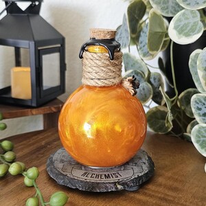 May include: A round, orange-colored glass bottle with a cork stopper and rope detailing. The bottle has a shiny, metallic finish and sits on a gray stone coaster with the word "ALCHEMIST" printed on it. A black lantern and greenery are in the background.