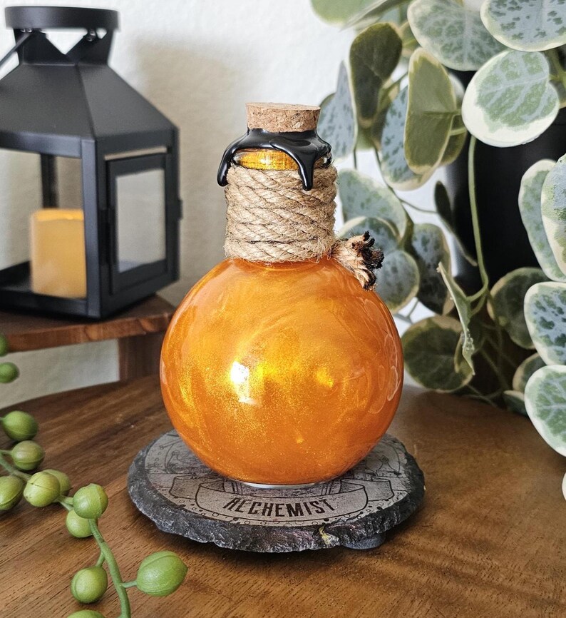 May include: A round, orange-colored glass bottle with a cork stopper and rope detailing. The bottle has a shiny, metallic finish and sits on a gray stone coaster with the word "ALCHEMIST" printed on it. A black lantern and greenery are in the background.