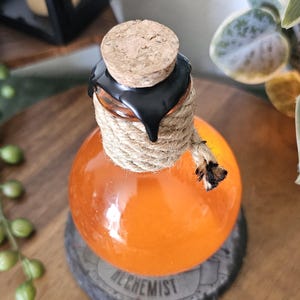 May include: Orange glass bottle with a cork stopper, black wax seal, and rope detailing. The bottle sits on a wooden coaster with the word "ALCHEMIST" printed on it. The bottle is filled with an orange liquid.