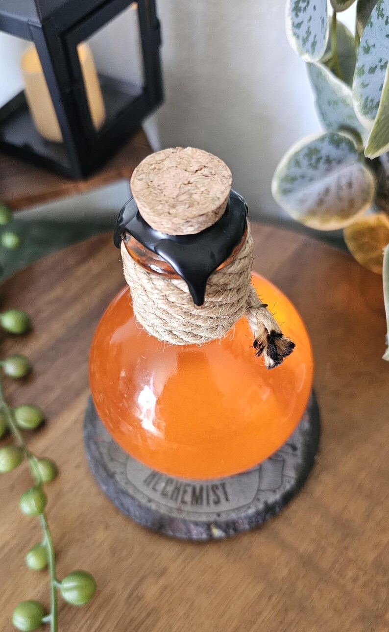 May include: Orange glass bottle with a cork stopper, black wax seal, and rope detailing. The bottle sits on a wooden coaster with the word "ALCHEMIST" printed on it. The bottle is filled with an orange liquid.