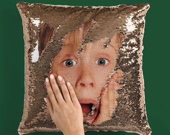 May include: A square sequined pillow with a gold-toned surface. The pillow features a printed image of a surprised face, partially revealed through a torn section of the sequins. A hand is touching the face.