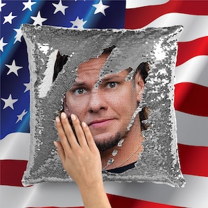 May include: A silver sequin pillow with a man's face peeking out from behind the sequins. The pillow is square and has a zipper closure.
