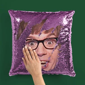 May include: A square, purple sequin cushion with a face peeking through a torn section. The face has glasses and an open mouth. A hand is touching the cushion.