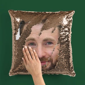May include: A square cushion with gold and brown sequins. The sequins reveal a face when brushed. A hand is touching the cushion. The background is a solid dark green.