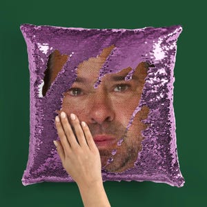 May include: A square decorative pillow with a reversible sequin design. The sequins are purple, revealing a man's face underneath. A hand is touching the pillow. The background is a solid dark green.